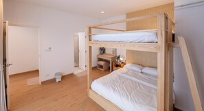 Habitación con literas, un escritorio, una silla, un cubo de basura pequeño y una puerta que da a otra habitación.
