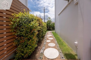 Un sendero estrecho con piedras redondas, bordeado por una cerca de madera y un seto, que conduce a un edificio blanco.