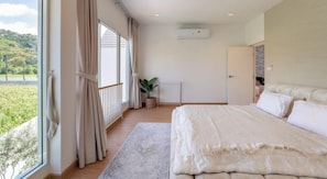 Een slaapkamer met een groot bed, uitzicht op groen en een raam met gordijnen.