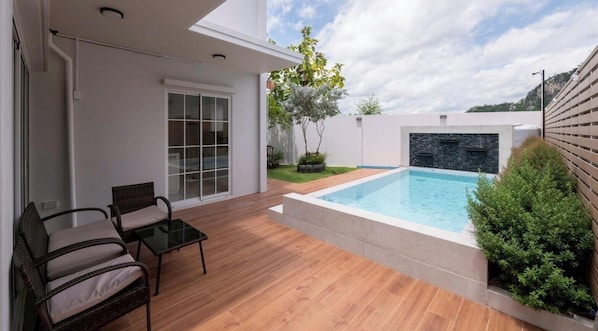 Ein moderner Poolbereich im Freien mit einer Holzterrasse, einem Glastür-Eingang und einer kleinen Sitzecke.