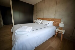 A neatly made bed with white linens and a folded towel, a wooden headboard, a bedside table with a lamp, and a wooden floor.