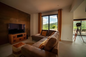 Een moderne woonkamer met een hoekbank, een tv op een houten dressoir en een groot raam met uitzicht op groen.