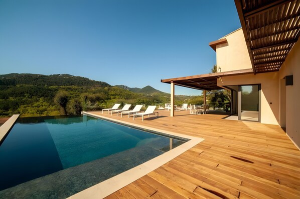 A modern pool area with wooden decking, lounge chairs, and a view of a mountainous landscape.