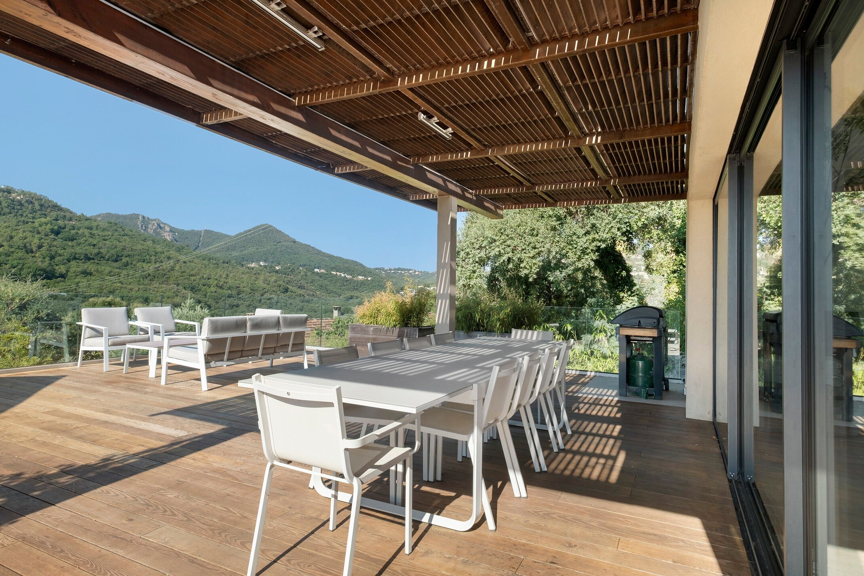 Een terras buiten met een houten pergola, een eettafel met stoelen en uitzicht op bergen.