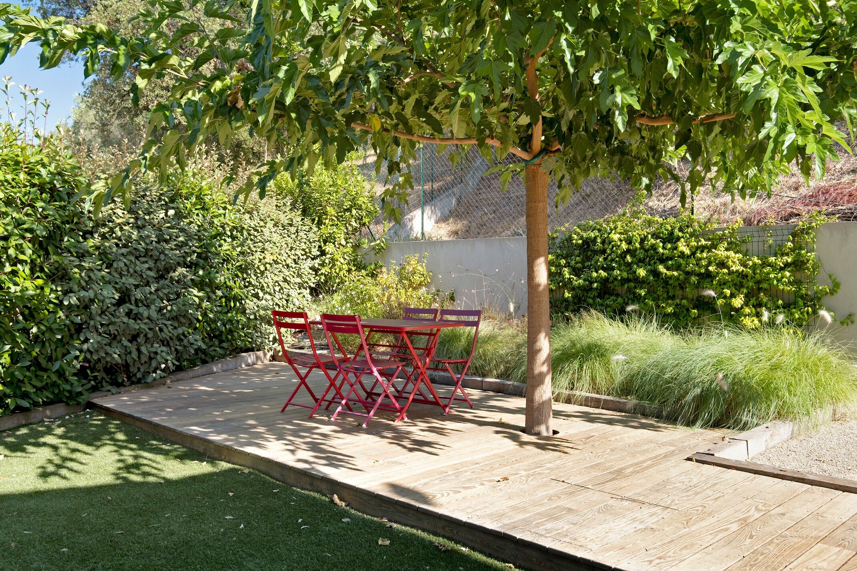 Une terrasse avec une table et des chaises, entourée de verdure et d’une clôture.