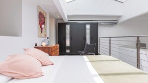 Un dormitorio moderno con tragaluz, una cama con almohadones rosados, un aparador de madera, una silla y un armario.