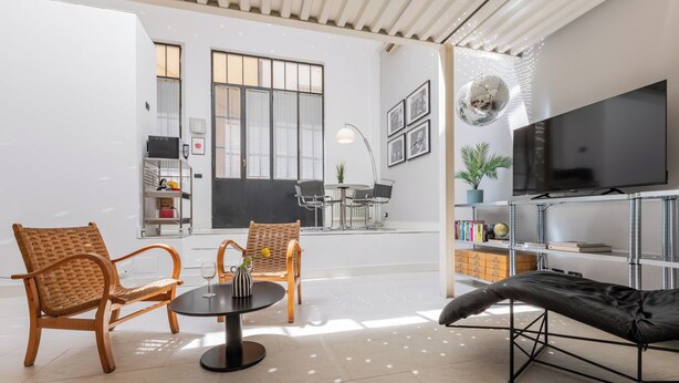 Una sala moderna con sillones de mimbre, un sofá negro, una mesa redonda y un televisor montado en un estante metálico.