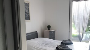 A bedroom with a bed, a small bedside table with a plant, a window with curtains, and a door.
