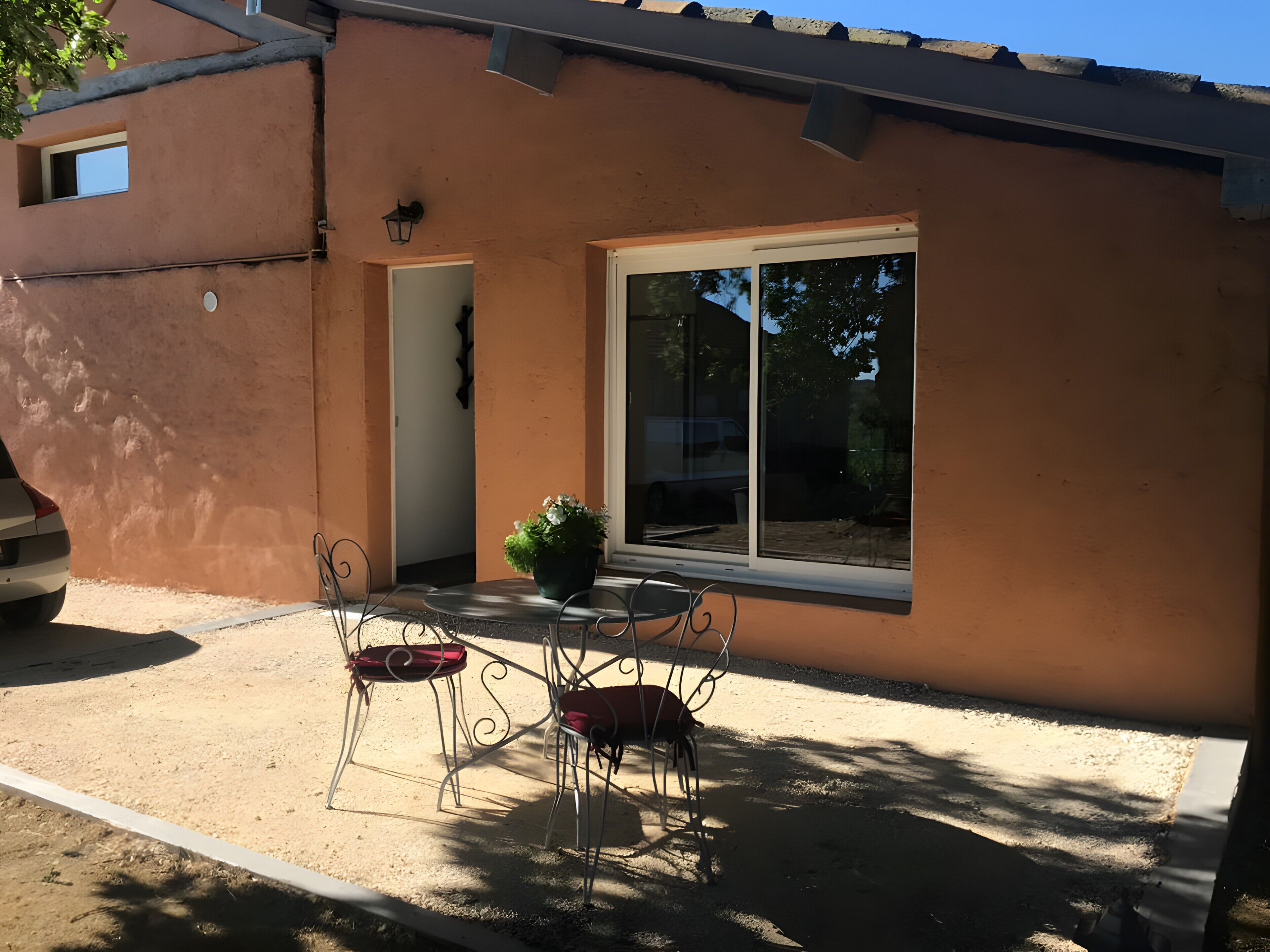 Un edificio de fachada marrón, un ventanal grande y un patio con sillas metálicas y una planta en maceta.