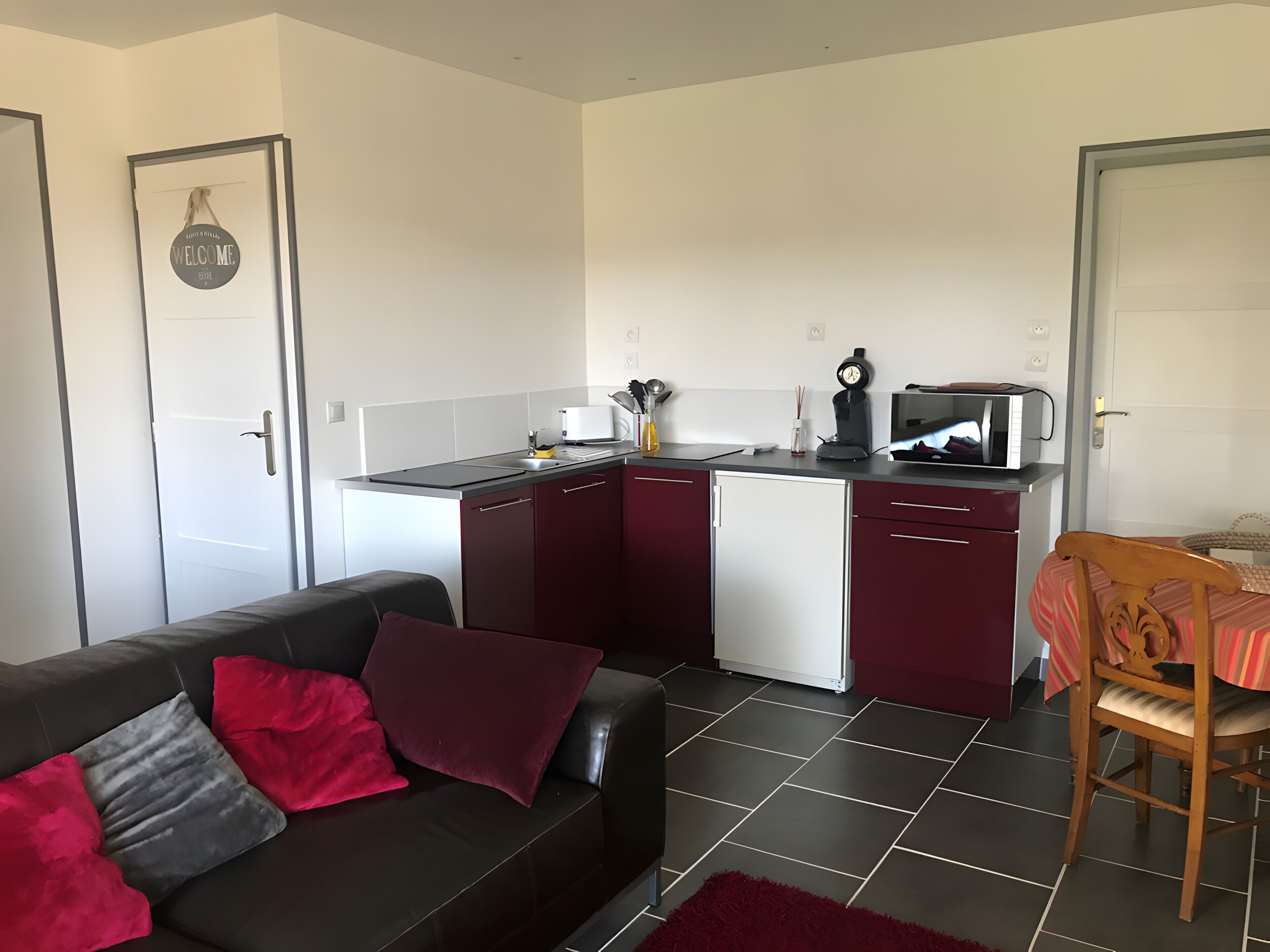 A compact kitchen with a microwave, toaster, and sink. There is a dining table with a chair and a sofa with red cushions.