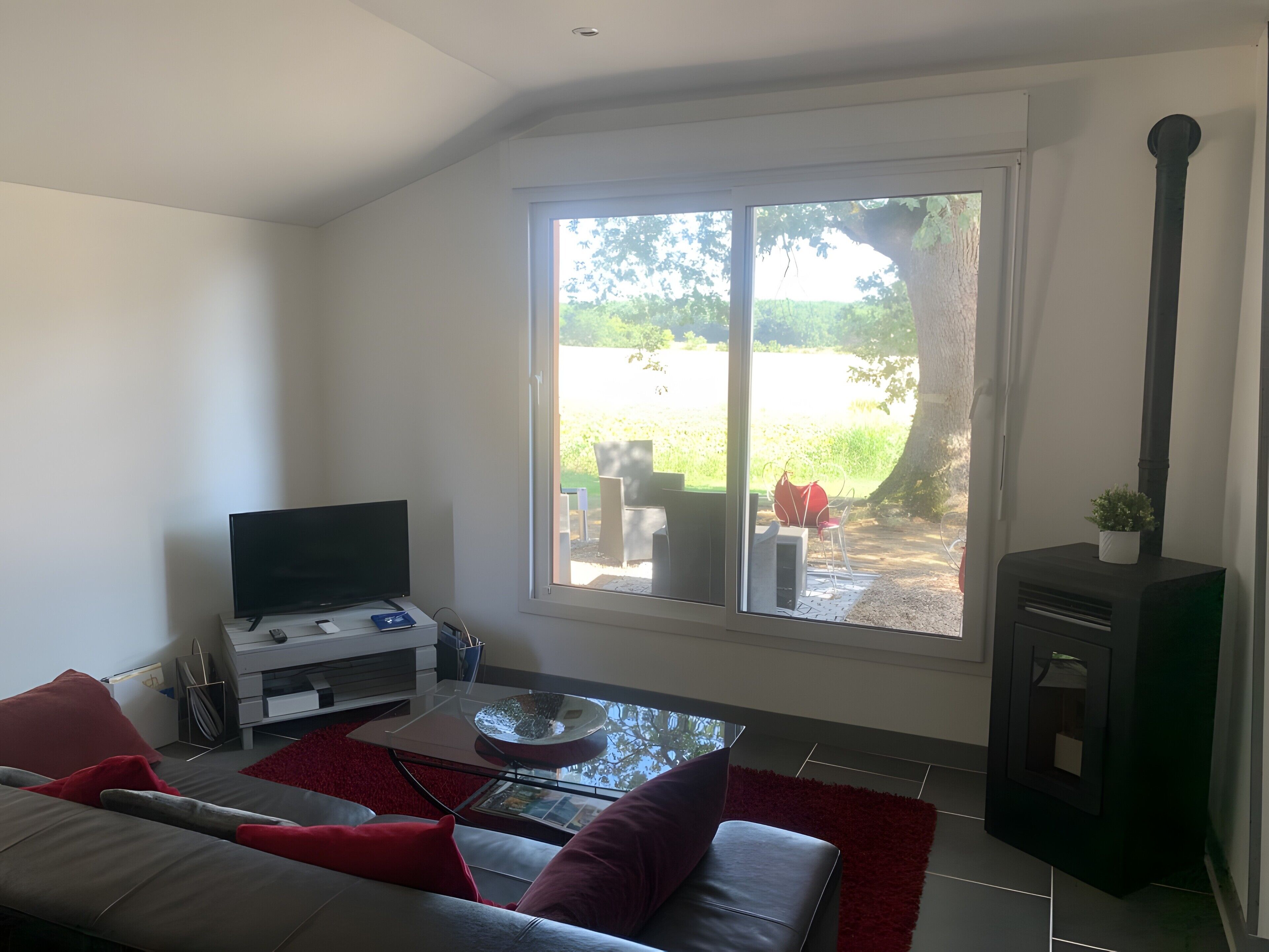 A modern living room with a flat-screen TV, a sofa with red cushions, a glass coffee table, and a large window offering a view of a garden.