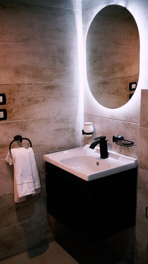 A modern bathroom with a large rectangular sink, a black faucet, a towel rack with a white towel, and a round mirror with a light. - Madina Boutique Hotel  (Luxor)