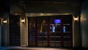 Entrada de edifício com porta de vidro grande, fachada de pedra e duas luzes fixadas na parede.