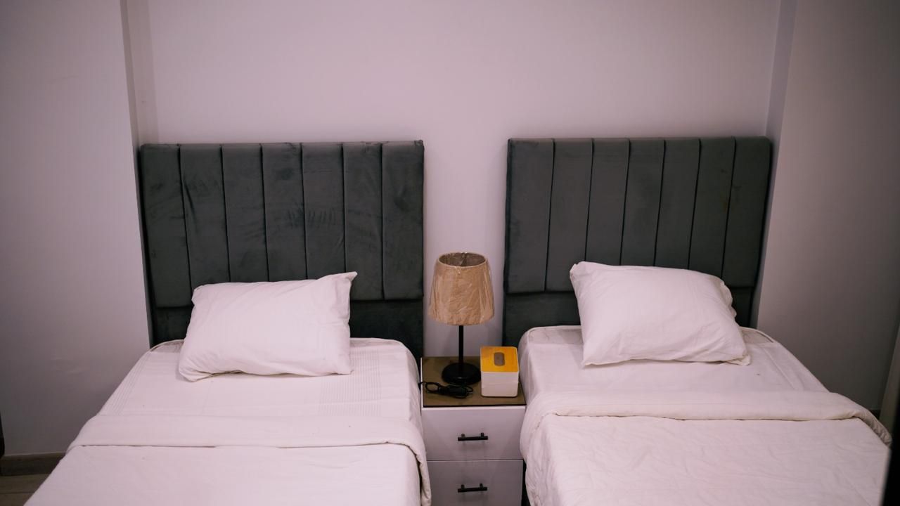 A room with two beds, each with a white pillow and a headboard. There is a nightstand with a lamp and a box between the beds.