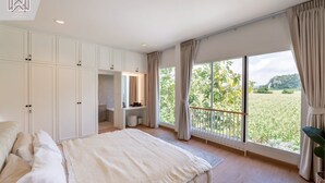 Ett sovrum med en stor säng, ett skrivbord och en garderob. Det finns en balkong med utsikt över grönområden och ett avlägset kull.