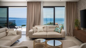 Ein modernes Wohnzimmer mit einer Couch, Sesseln und einem Couchtisch, mit großen Fenstern und Meerblick.