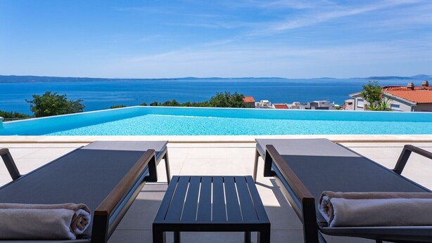 Ein Poolbereich mit Liegestühlen und einem Tisch, mit Blick auf das klare, blaue Meer und entferntes Land.