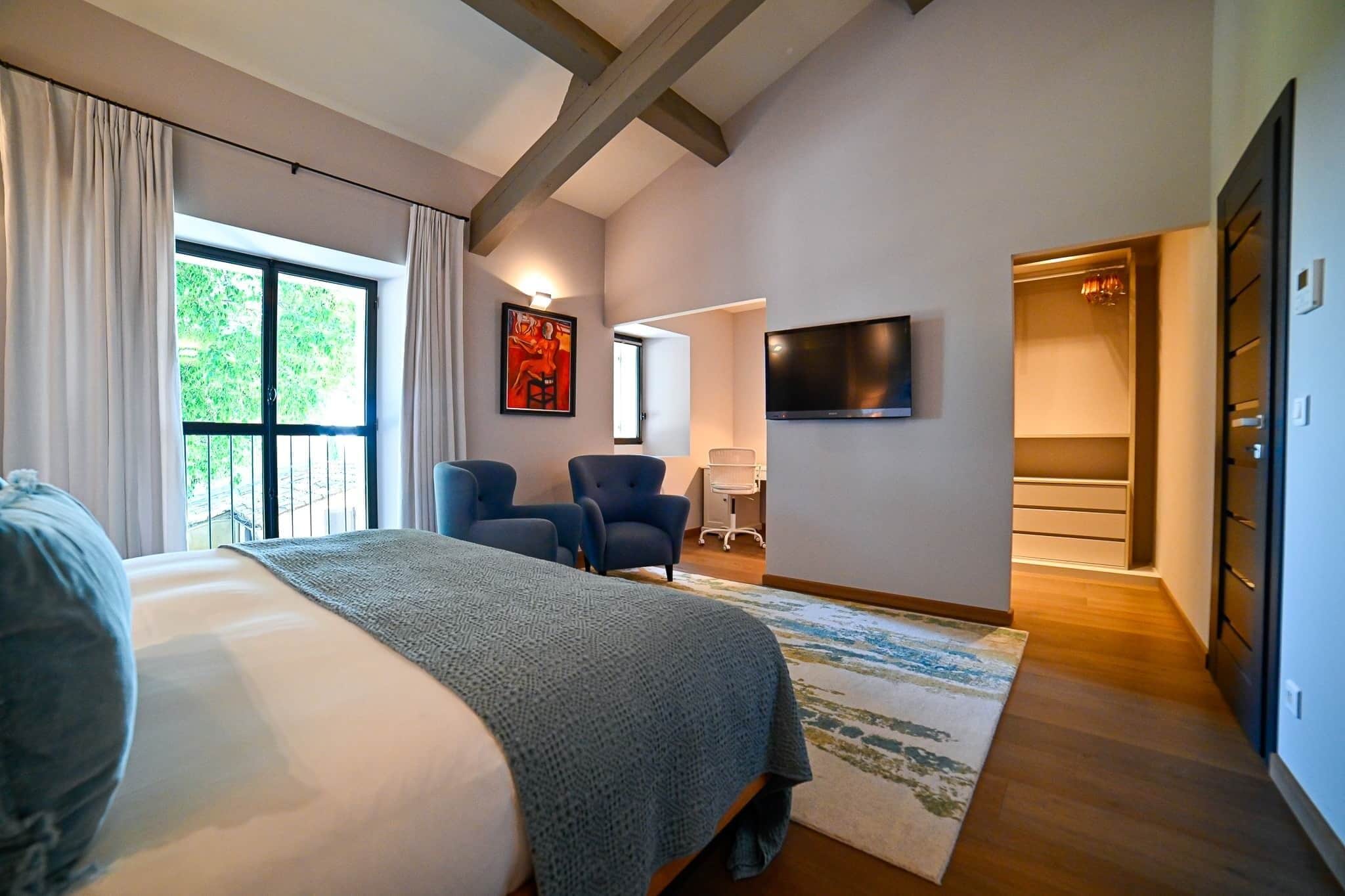 A bedroom with a large bed, two chairs, a TV, and a view of the outdoors.