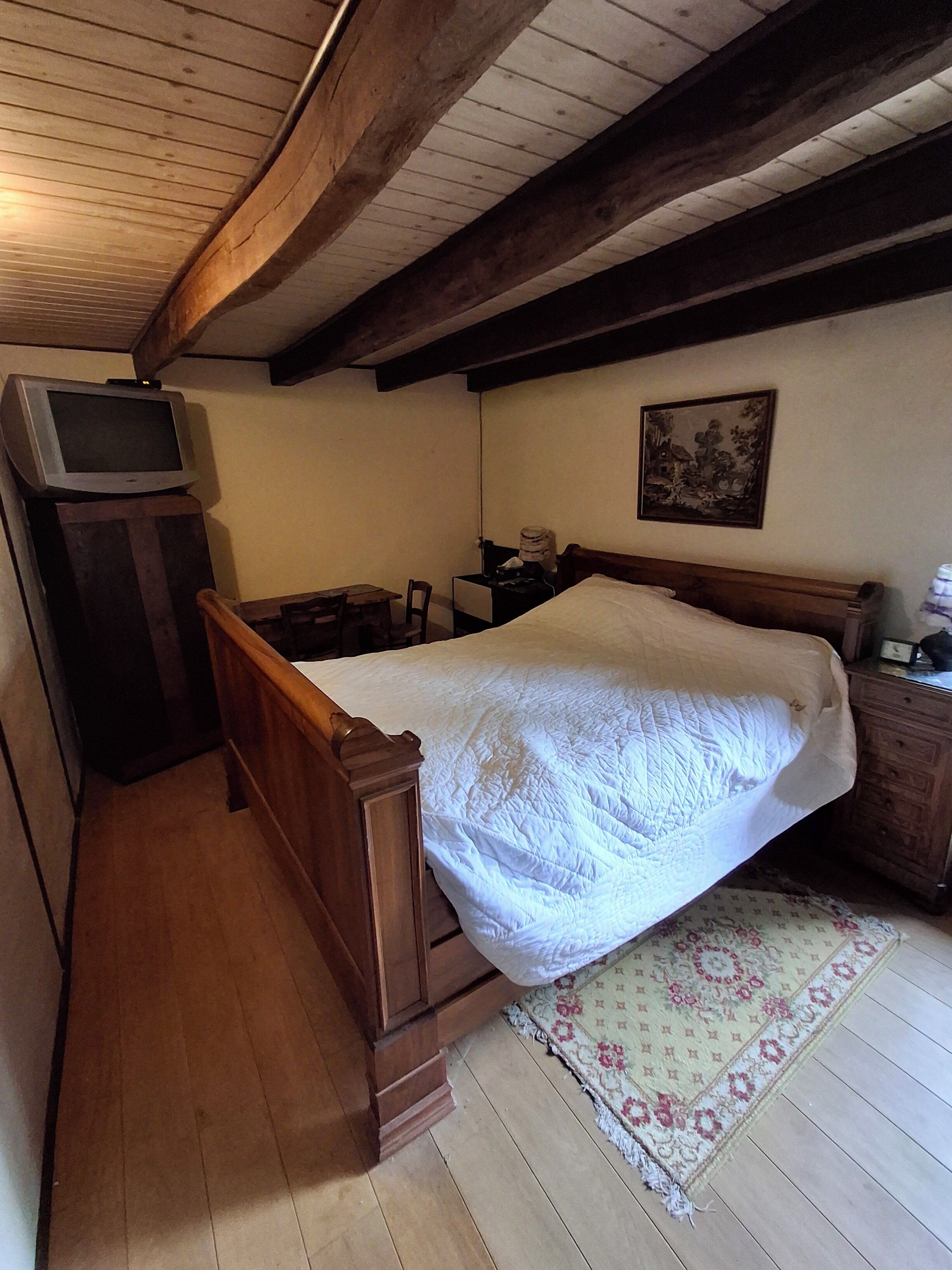 Une chambre à coucher avec un lit en bois, un téléviseur posé sur un meuble en bois, une table de chevet, une chaise, un tapis et un tableau accroché au mur.