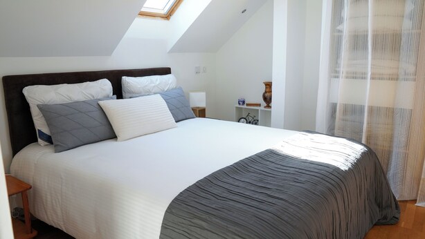 Un dormitorio con tragaluz, cama con ropa de cama blanca y gris, mesita de noche y un pequeño adorno.