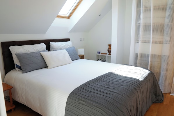 A bedroom with a skylight, a bed with white and grey bedding, a bedside table, a nightstand, and a small decorative item.