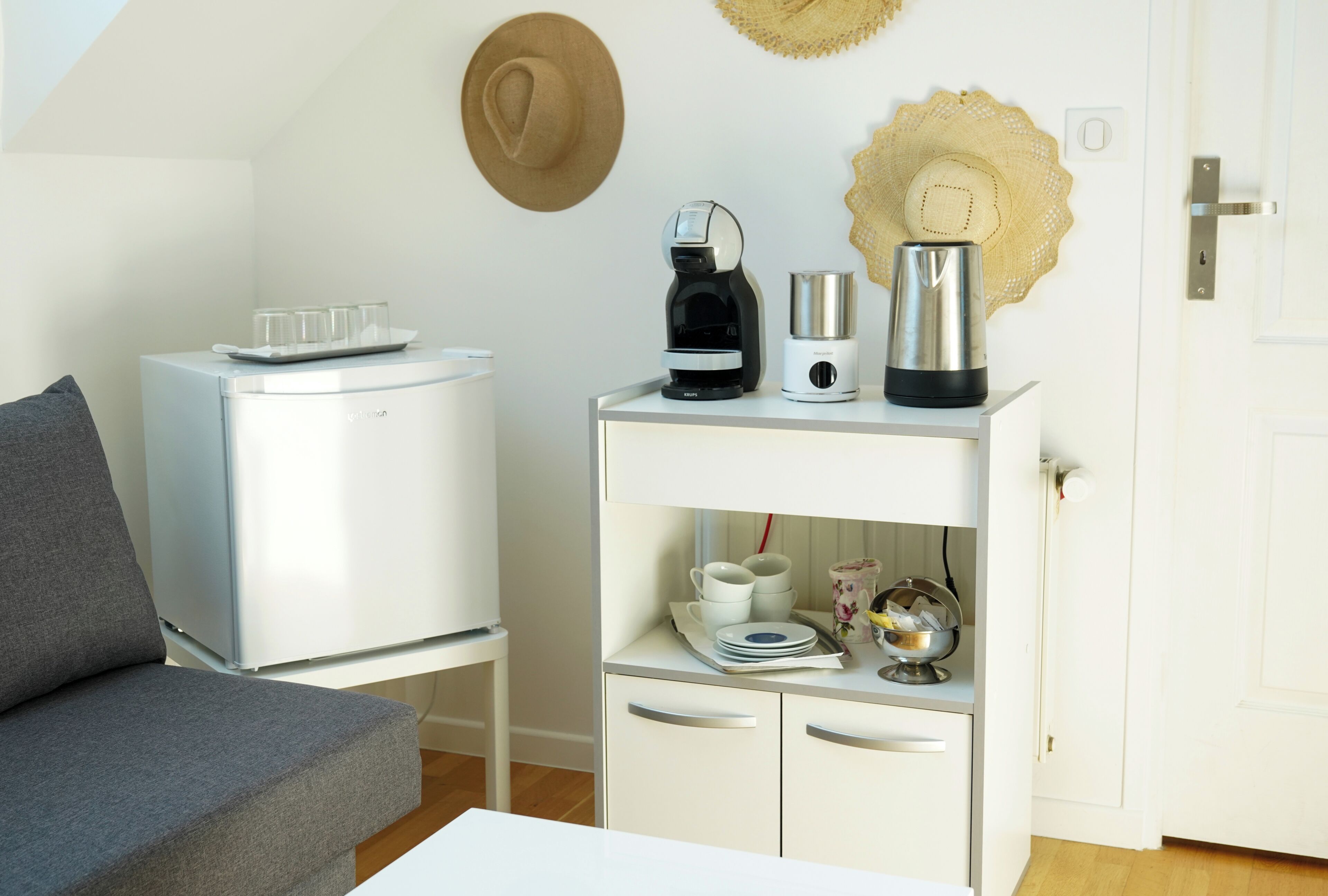 Un espace de vie compact comprenant un canapé, un petit meuble blanc contenant des appareils de cuisine, et une porte dotée d’une poignée.