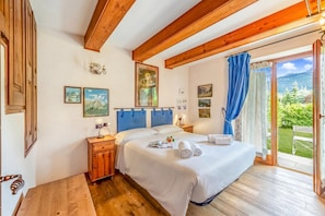 A bedroom with a wooden ceiling, a bed with white linens, a nightstand, a small table, and a view of greenery through a sliding door.