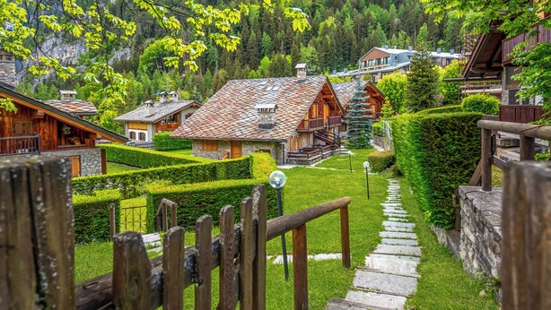 A wooden cabin with a stone pathway leading to a wooden deck, surrounded by lush greenery and a mountainous backdrop.
