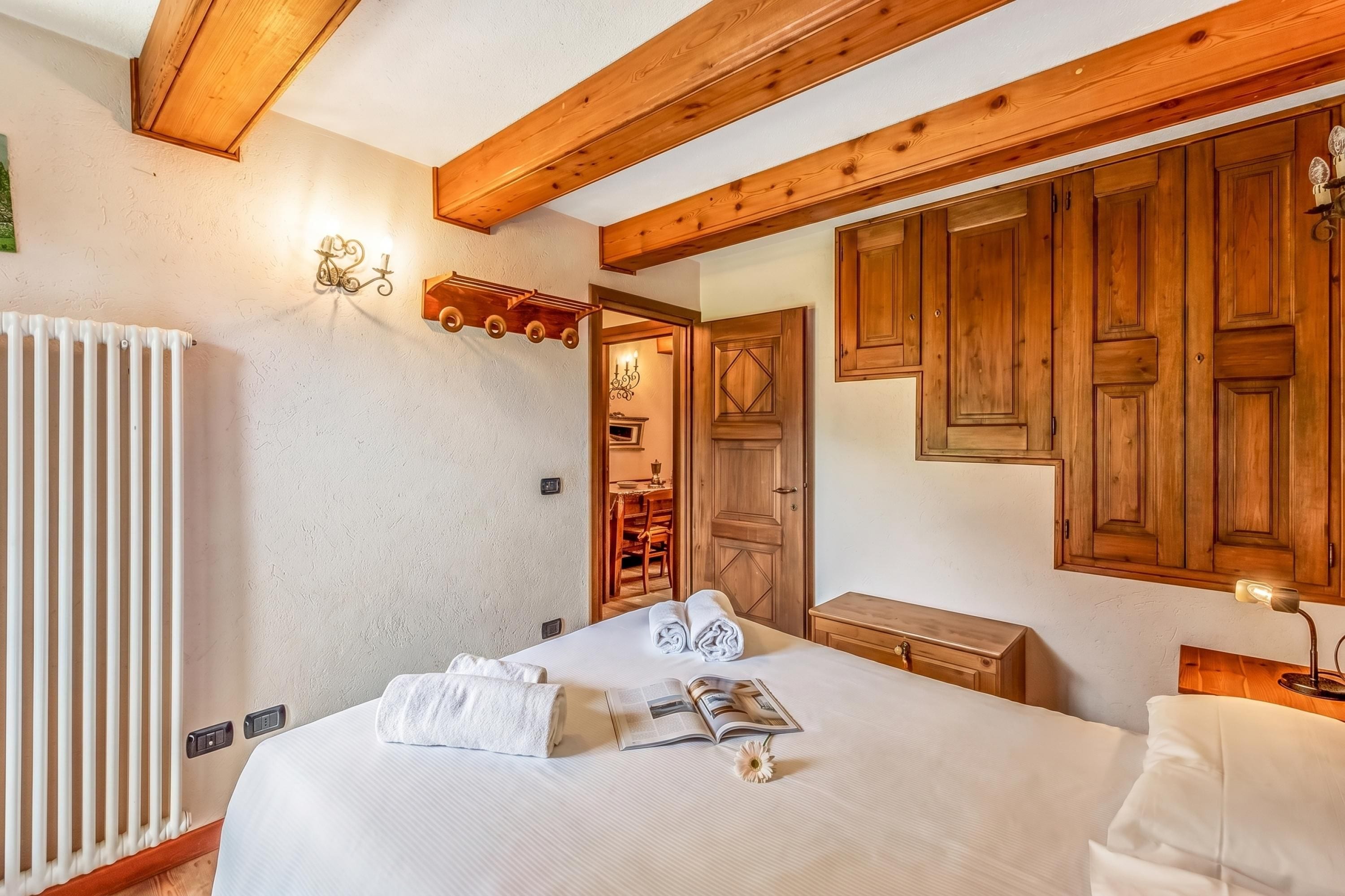 A bedroom with a bed, wooden ceiling beams, a radiator, a wall-mounted light fixture, and a wooden wardrobe.