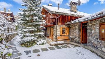 Un edificio de madera cubierto de nieve con muros de piedra, una escalera de piedra y un árbol nevado.