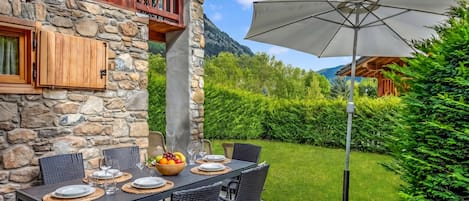 An outdoor dining area with stone walls, a table set with plates, glasses, and a fruit bowl, and a large umbrella.