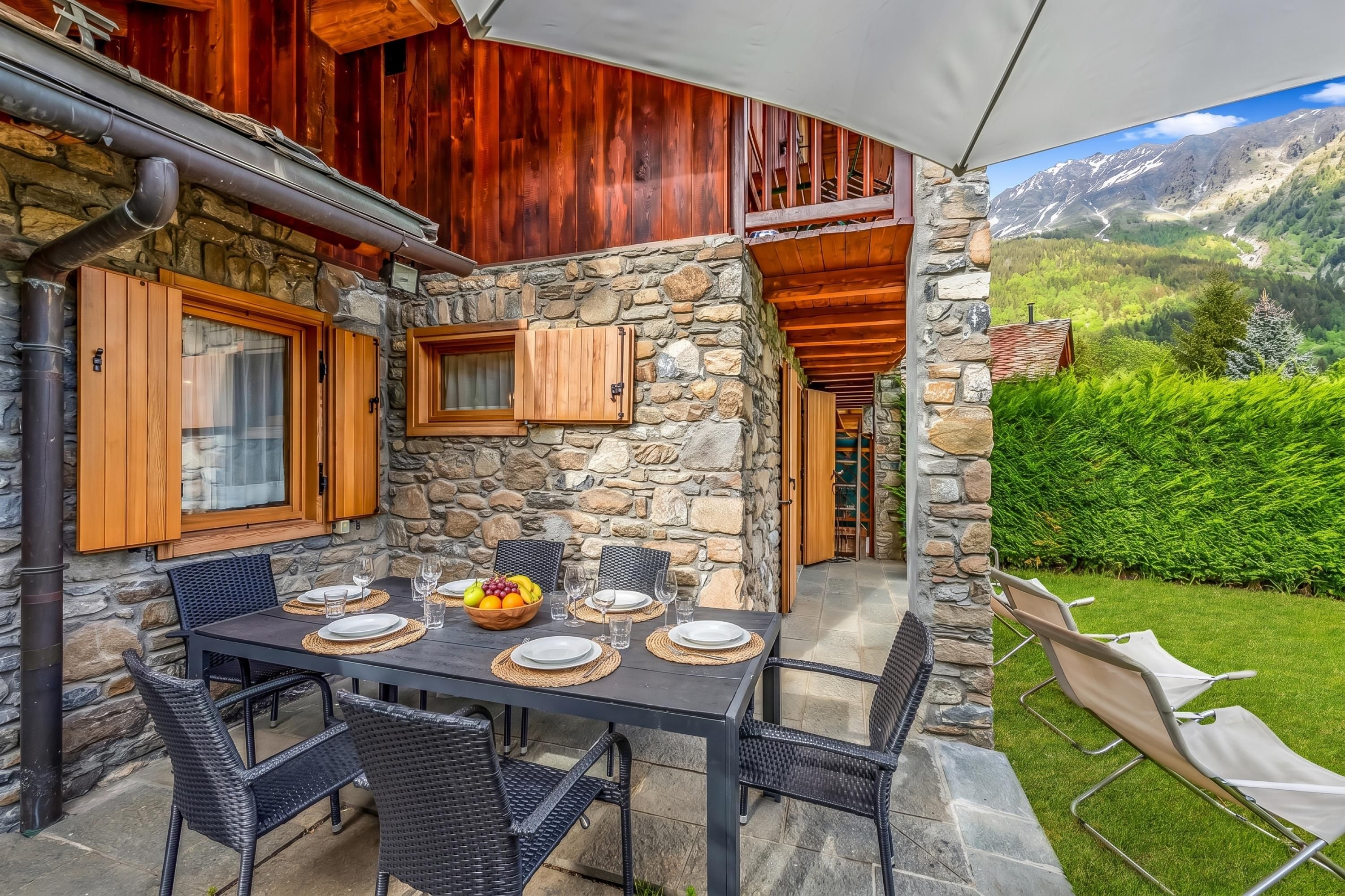 A stone exterior with wooden shutters, a dining table set with plates, glasses, and a fruit bowl, and a view of mountains.