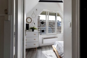 A bedroom with a bed, a dresser, a radiator, and a window with a view of buildings. - Holiday Home 'Maison De Molène' with Sea View (Trébeurden)