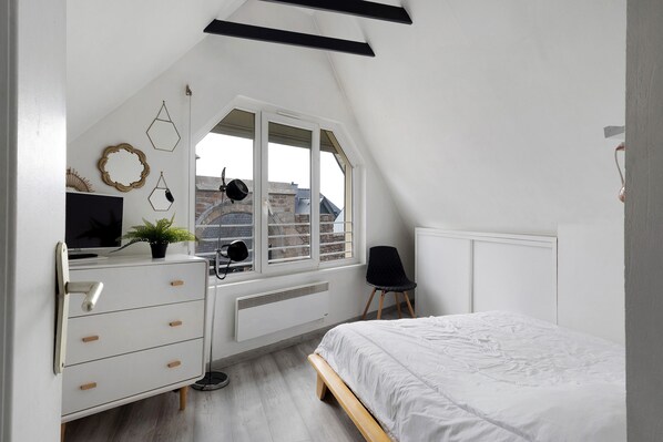 Ein Schlafzimmer mit schrägen Wänden, einem Schreibtisch mit Computer, einem Stuhl, einem Bett und einem Fenster mit Blick auf Gebäude.