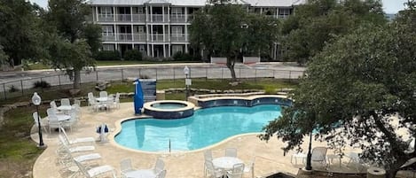 Piscina com espreguiçadeiras e hidromassagem, cercada por uma área delimitada e um edifício ao fundo.