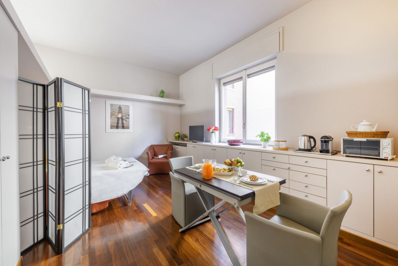 A compact room with a bed, a dining table with breakfast, a kitchenette, and a large window.