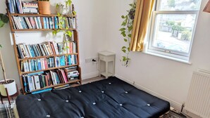 Een kamer met een zwart matras, een boekenplank vol boeken, een potplant en een raam met gordijnen.