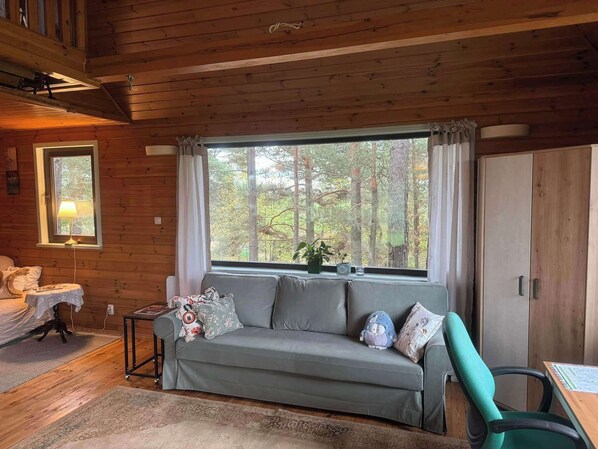 Ein gemütliches Blockhaus-Interieur mit einer Couch, einem Tisch, einer Lampe und Blick auf einen Wald durch ein Fenster.