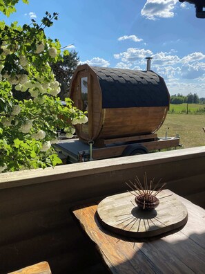 A wooden barrel-shaped structure with a chimney, a wooden table with a decorative object, and a view of a green field. - Cosy Cabin with Fireplace in Podlasie (Czarna Wieś Kościelna)