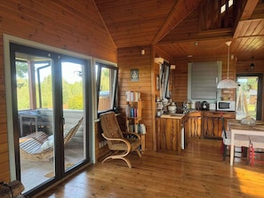 A wooden cabin interior with a dining area, kitchen, and a large sliding glass door leading to an outdoor deck.