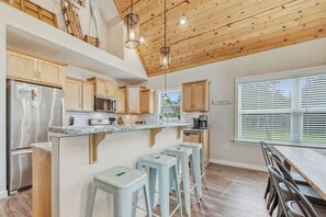 Eine Küche mit Holzausstattung, einem Edelstahlkühlschrank, einer integrierten Kücheninsel mit Hockern und einem Fenster mit Jalousien.
