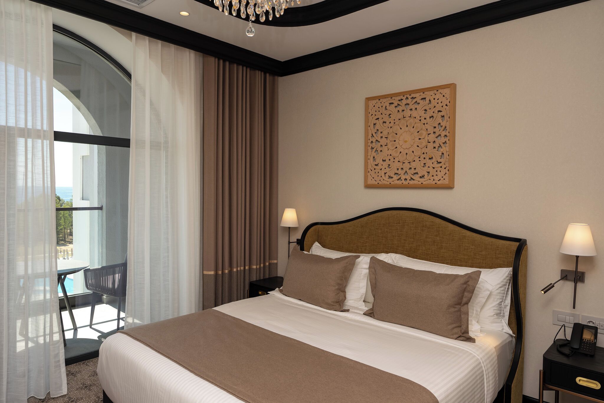 A hotel room with a bed, two bedside lamps, a decorative wall panel, a window with curtains, and a view of the outdoors.