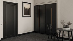 Una habitación con una puerta oscura, un cuadro en la pared, un armario negro, una mesa redonda con un jarrón y una silla.