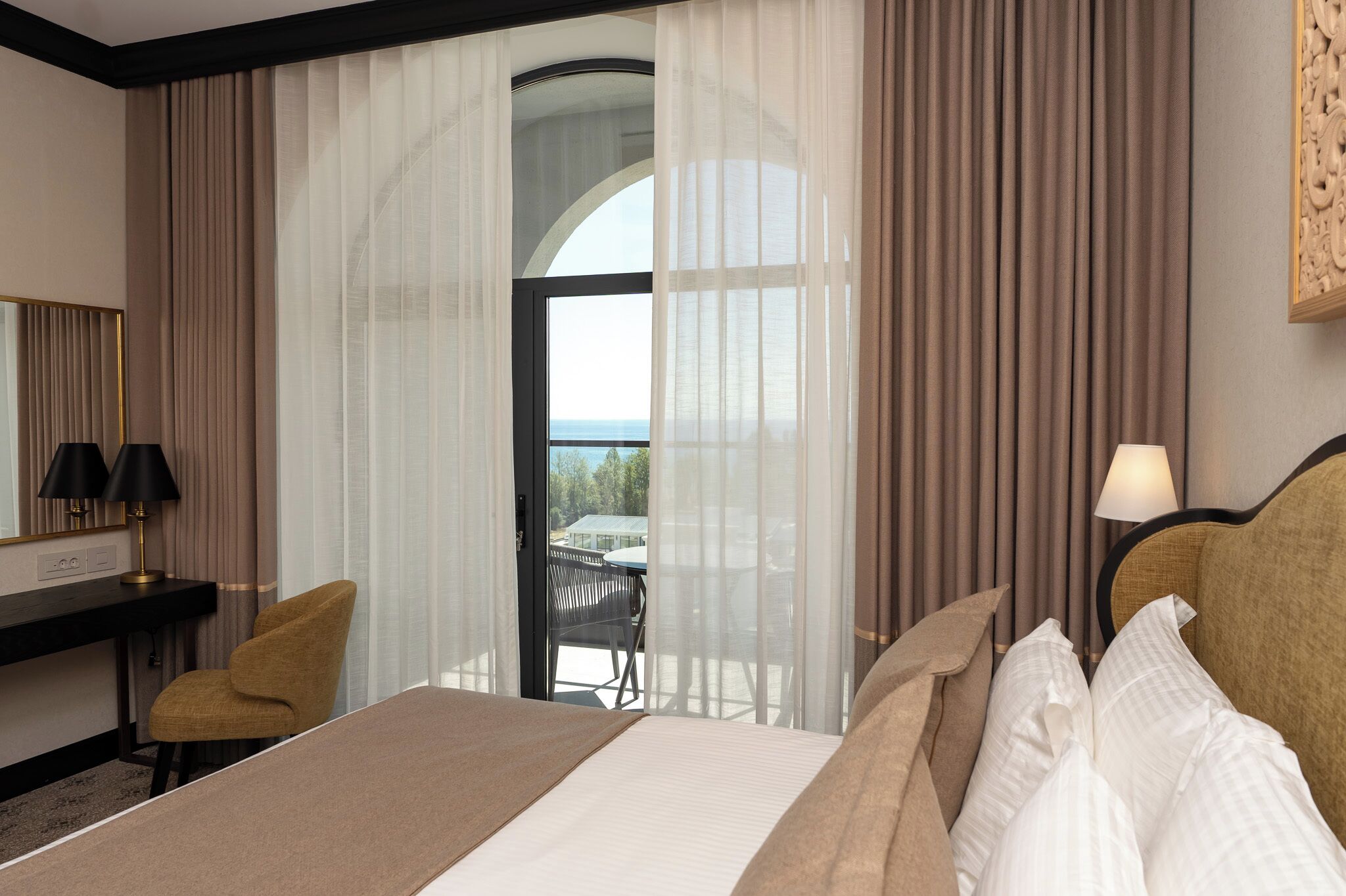 Habitación de hotel con cama, escritorio con lámparas, una silla y vista al exterior a través de un ventanal grande.
