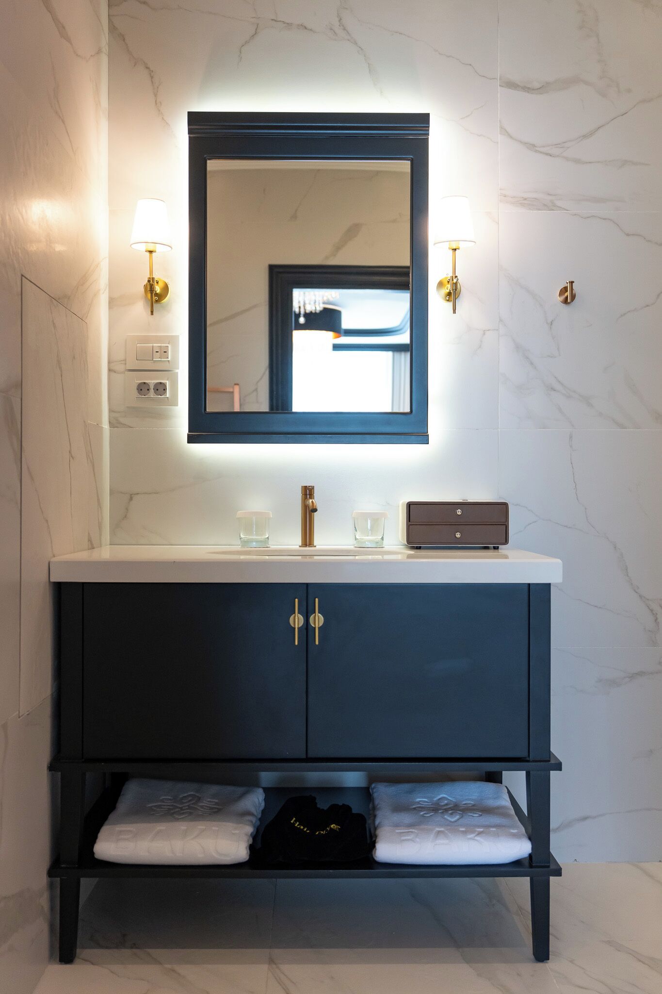A modern bathroom with a marble wall, a large mirror, two wall-mounted lights, a sink with a faucet, and a cabinet with two doors.