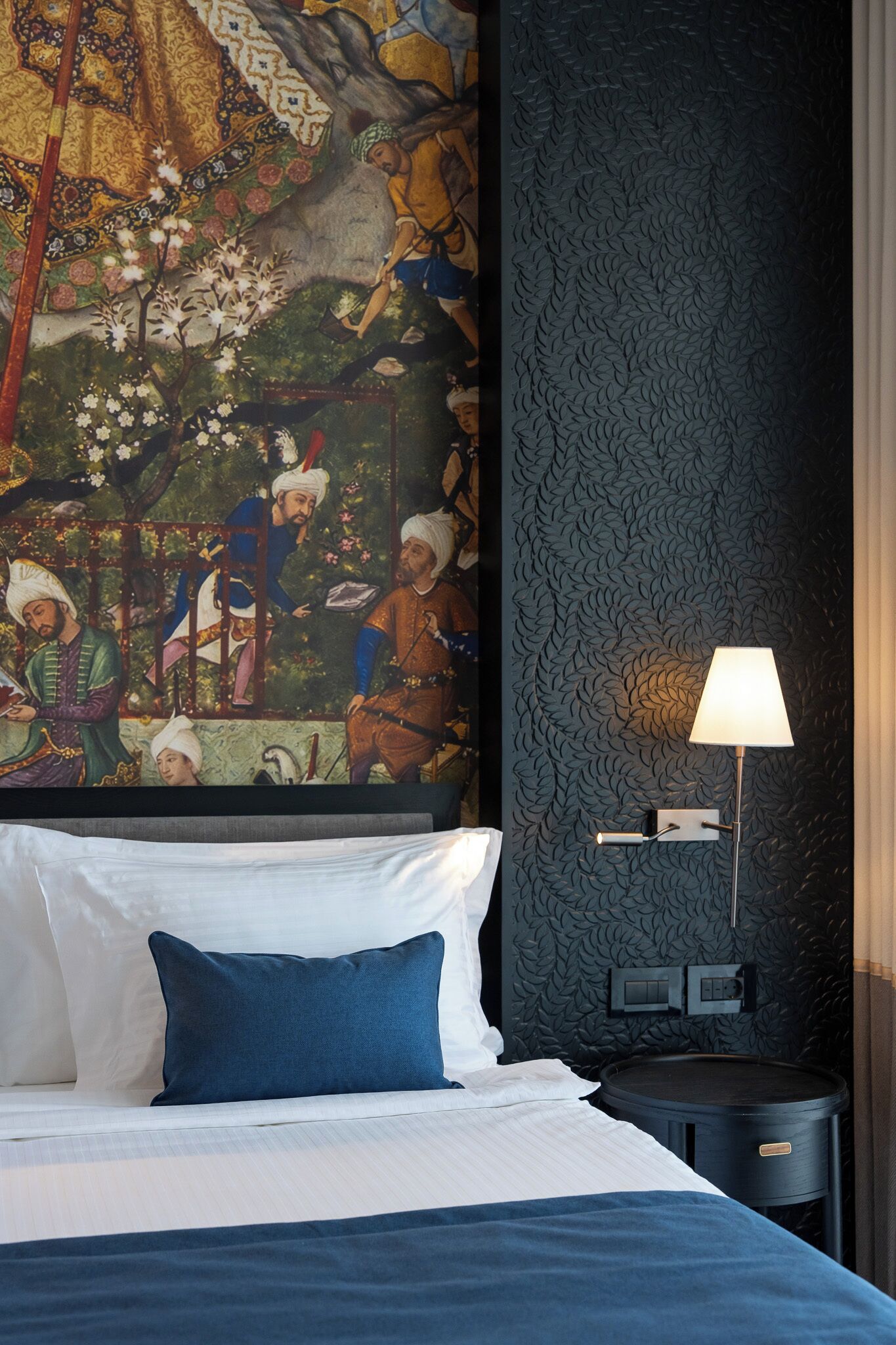 Habitación de hotel con un gran mural en la pared, una cama con ropa de cama blanca y azul, una lámpara y una mesita de noche pequeña.