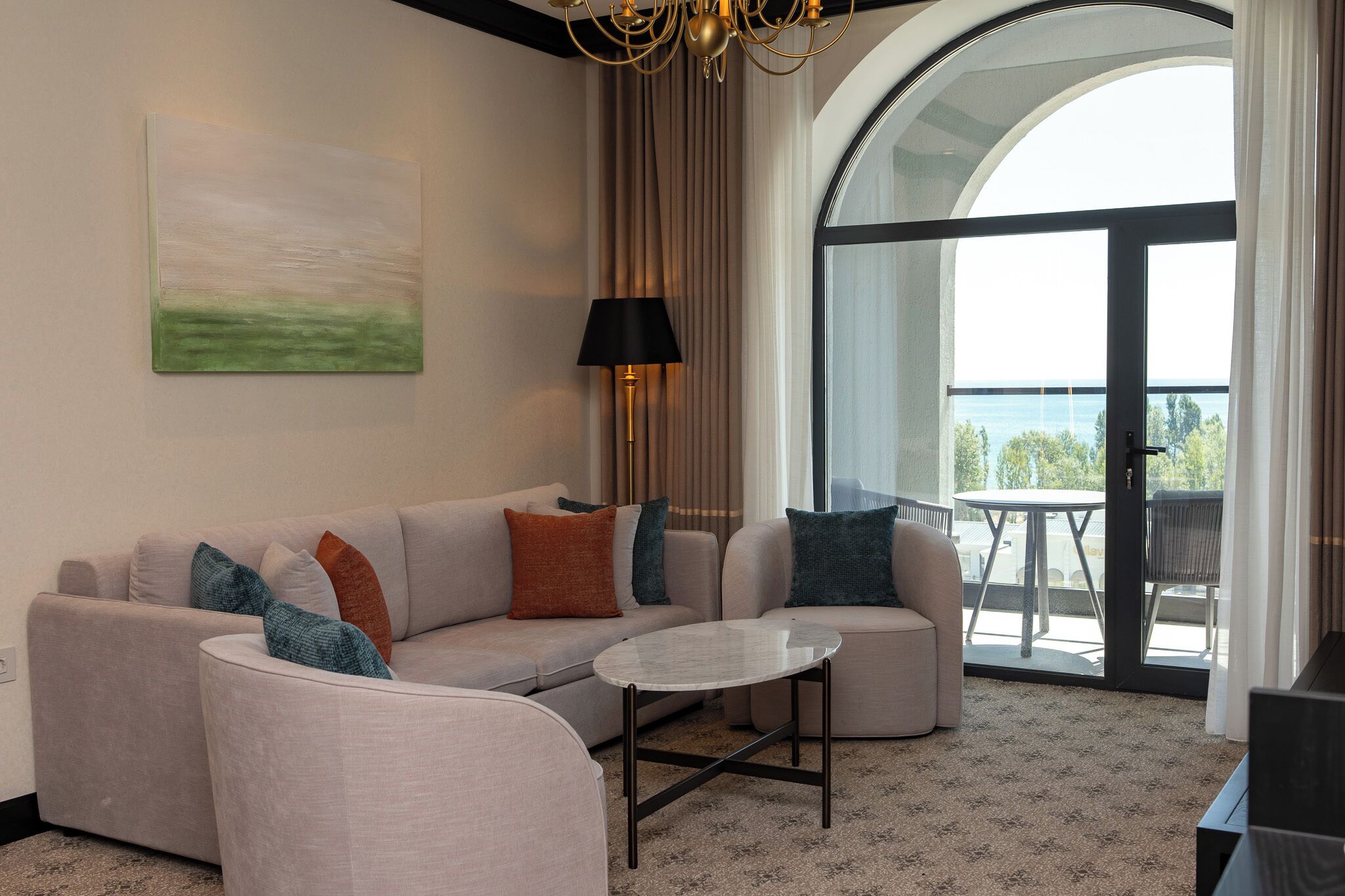 A modern living room with a sofa, a round coffee table, and a lamp. There is a large window with a view of the outdoors.