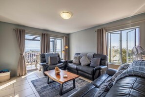 A modern living room with a sofa, a coffee table, and a view through large windows. - Charming 2-bedroom Apartment With Ocean and Mountain Views (Plettenberg Bay)