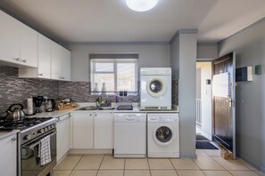 A compact kitchen with a stove, oven, washing machine, and a window with blinds. - Charming 2-bedroom Apartment With Ocean and Mountain Views (Plettenberg Bay)
