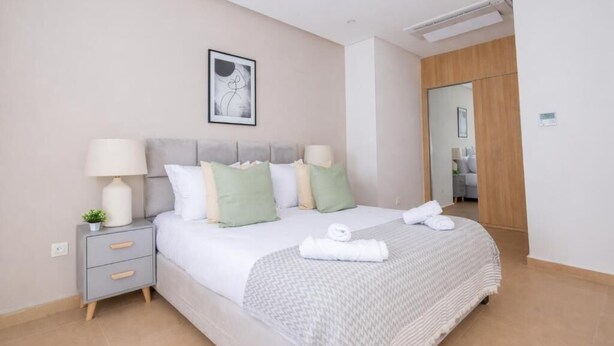 A neatly made bed with a grey headboard, a bedside table with a lamp, and a framed picture on the wall. - Appartement Charmant a hydra (Algiers)